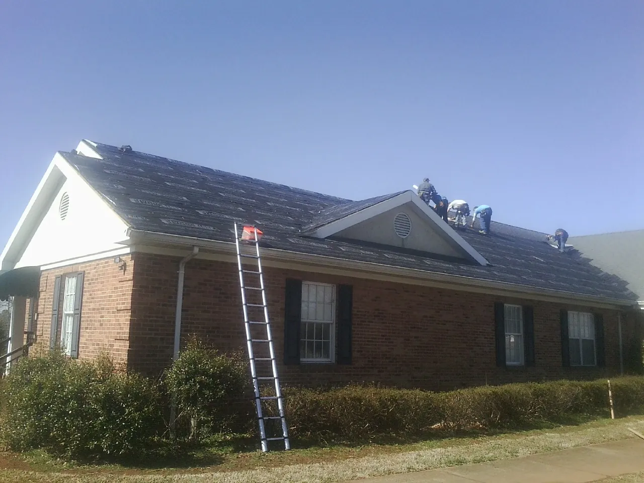 Crew tearing shingles off a brick ranch roof with a ladder set — Emergency Roof Repair work by our crew in Fishers