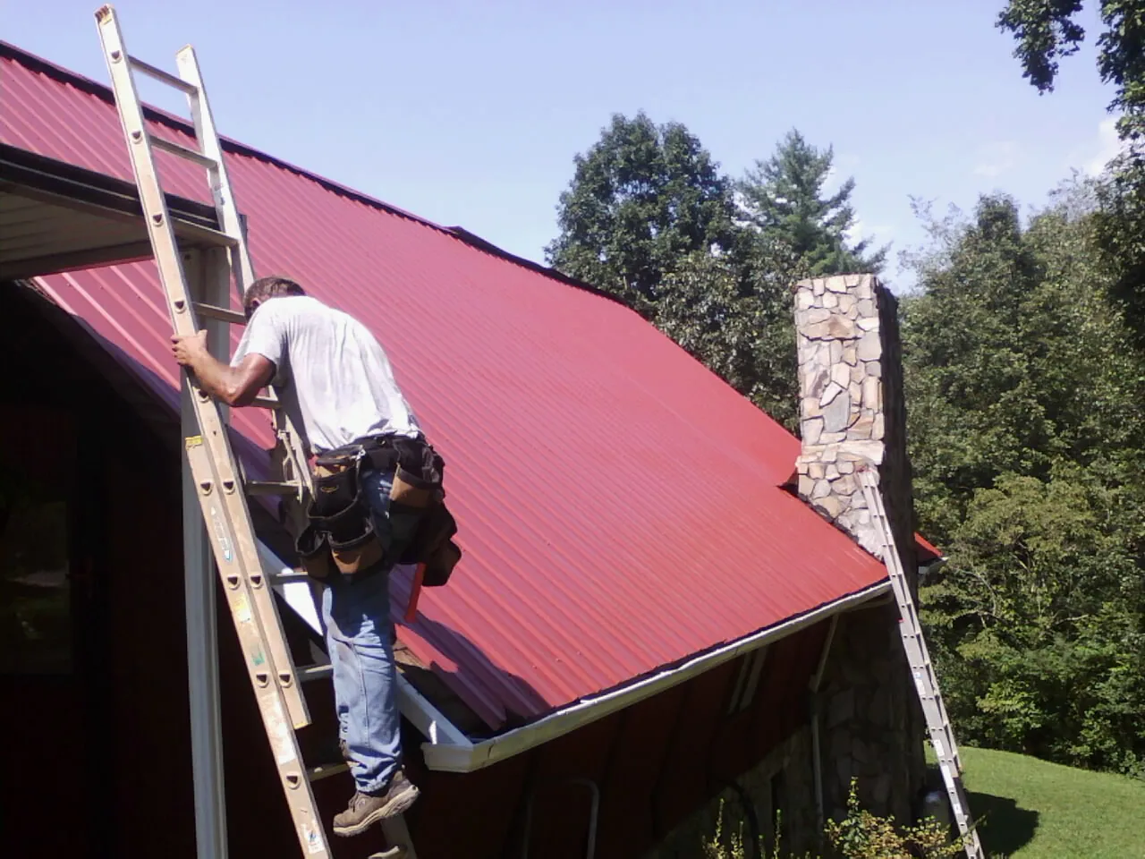 Roofer on a ladder beside a red metal roof and stone chimney — Soffit & Fascia Repair by Roofing Brothers in Fishers
